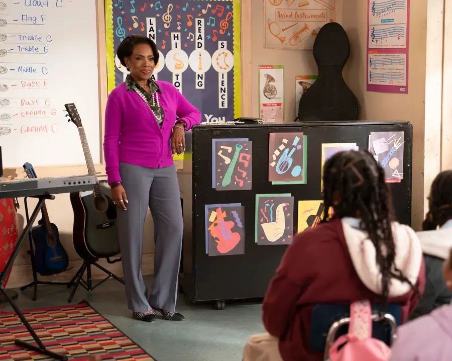 A teacher in a purple cardigan stands by a decorated bulletin board in a music classroom. Students sit facing her. Musical art decorates the room.