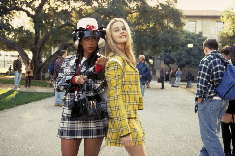 Two women in plaid outfits, one in yellow, the other in black and white, talk in a sunny school courtyard. People and trees in the background.