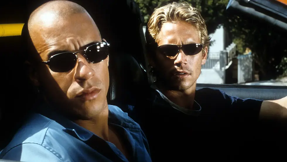 Two men in sunglasses sit in a car, staring ahead with serious expressions in bright daylight.