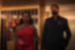 A woman in a red dress and a man in a dark suit stand elegantly in a room with floral decorations, exuding confidence and sophistication.