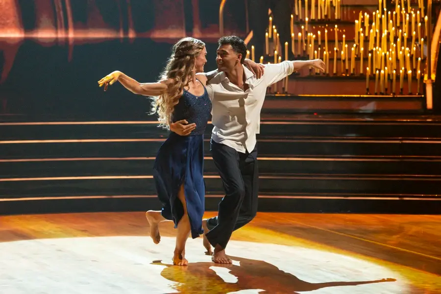 Dancing couple in navy and white on a lit stage with candlelit backdrop, arms extended in a graceful, intimate pose.