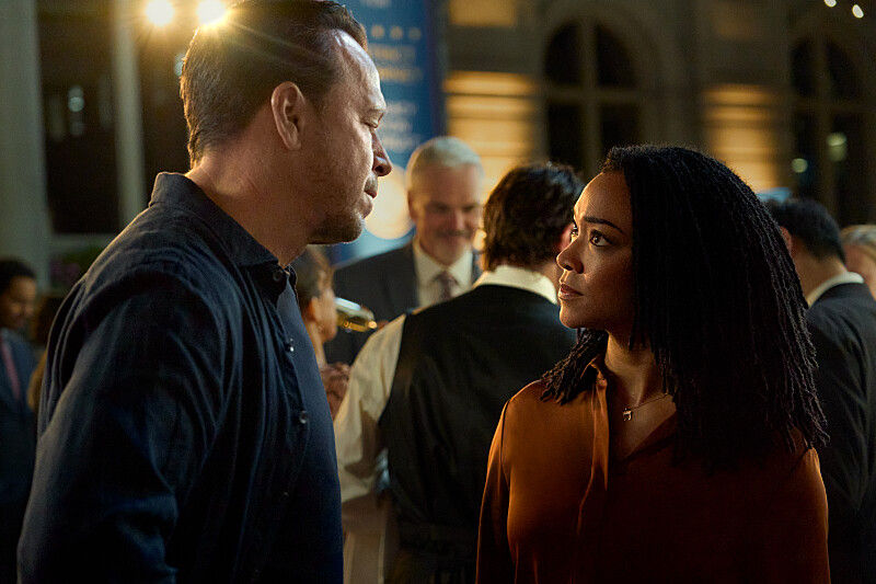 Man and woman face off at a warm-lit party, with blurred guests in the background and a tense mood.