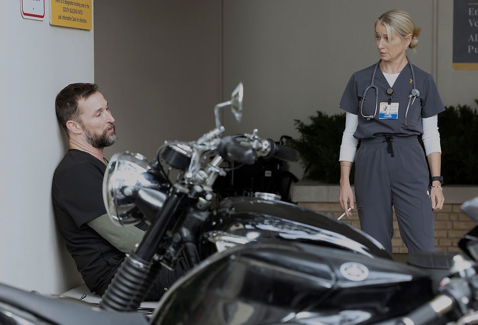 A man sits by parked motorcycles, appearing thoughtful. A nurse stands nearby, looking at him. Background includes greenery and signage.