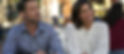 A man in a blue shirt and a woman in a white polka dot blouse sit outside, looking thoughtful and content. Blurred background with people.
