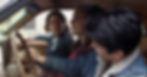 Three people sit in an old car, the driver glancing back at two passengers in a tense conversation outside a house.