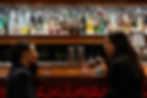 Two women chat at a warmly lit bar, holding drinks. A colorful array of bottles lines the shelves in the background.