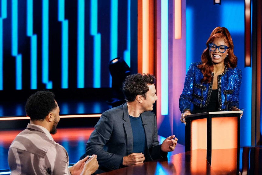 Woman in blue sequined jacket smiles at podium on a colorful game show set while two men watch.