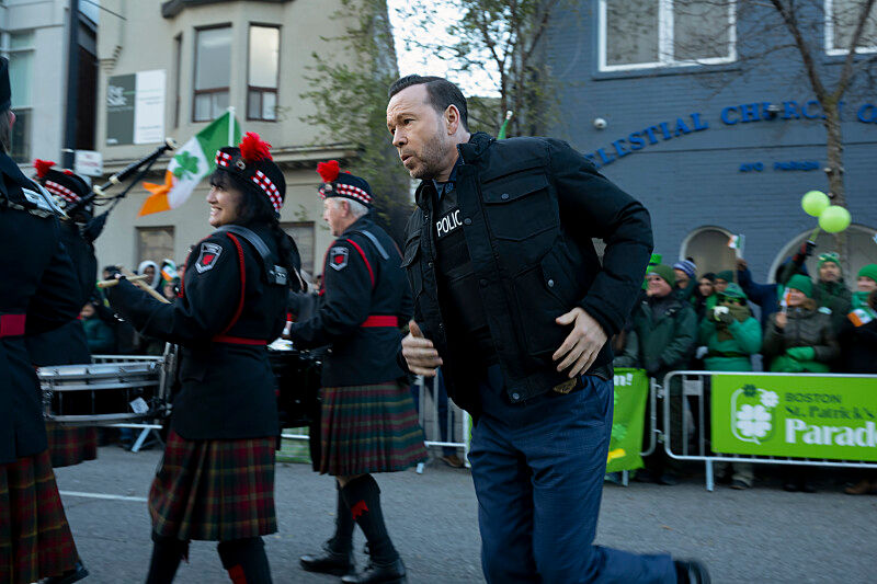 A man in a police vest runs alongside a marching band in kilts during a St. Patrick's Day Parade, with spectators and Irish flags in the background.