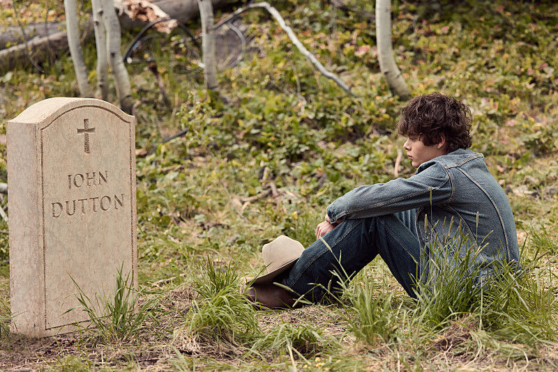 Boy in denim sits beside a gravestone marked JOHN DUTTON in a grassy wooded area, looking somber.