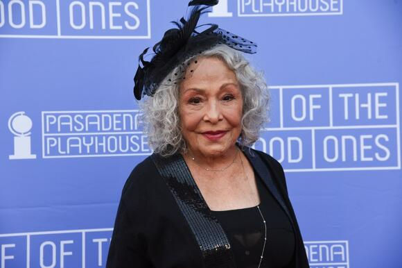 Elderly woman with curly gray hair and black outfit smiles at camera. Blue background with "PASADENA PLAYHOUSE" text. Elegant mood.