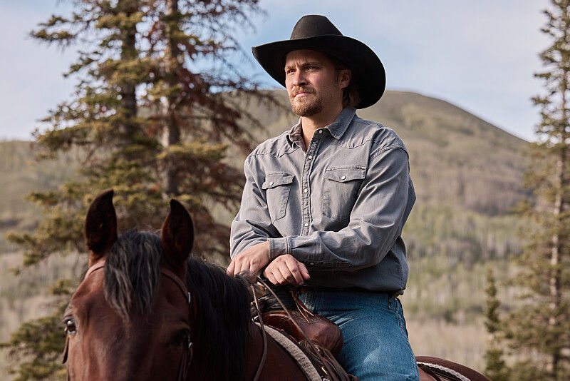 Cowboy in black hat rides a brown horse in a mountain forest, looking serious against pine trees and hills.