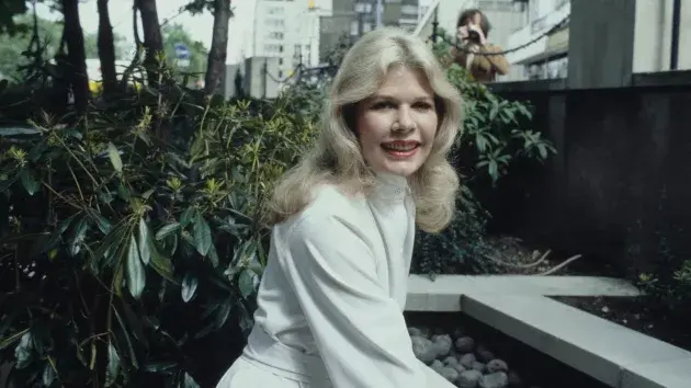 Woman in a white outfit smiles, sitting outdoors by greenery and a stone path. Urban background with trees and buildings.