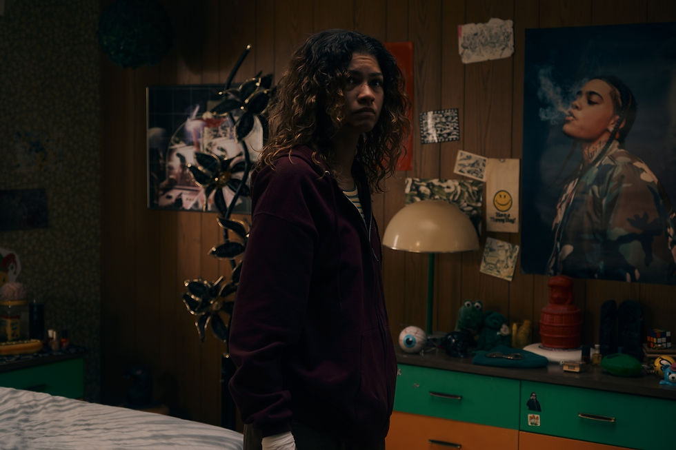 Teen girl in a dim bedroom, wearing a burgundy hoodie, stands by a poster-covered wall with a moody, uneasy expression.