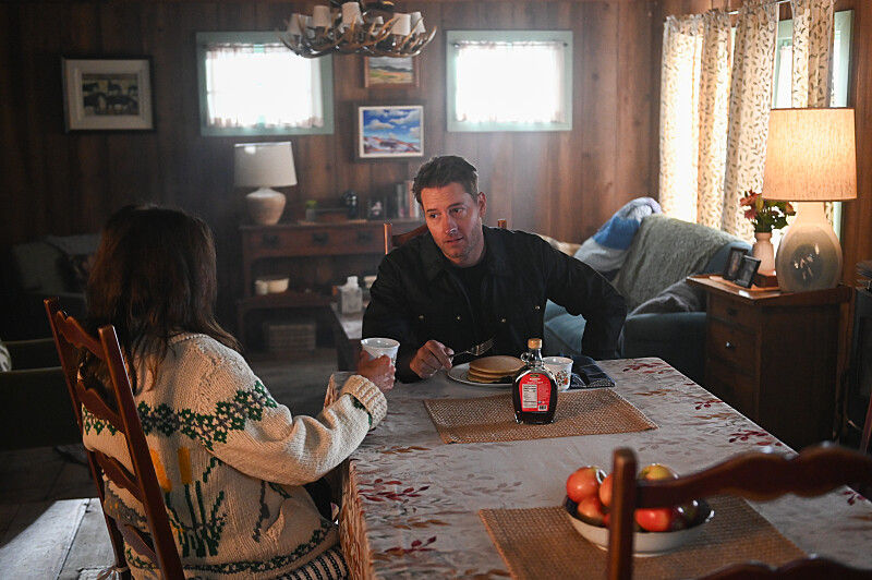 Two people sit at a dining table in a cozy, wood-paneled room. One wears a patterned sweater. Pancakes and syrup are on the table. Warm lighting.