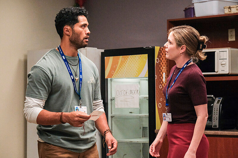 Two people with ID lanyards talk intensely in an office break room. A fridge with "Out of Order" sign is in the background.