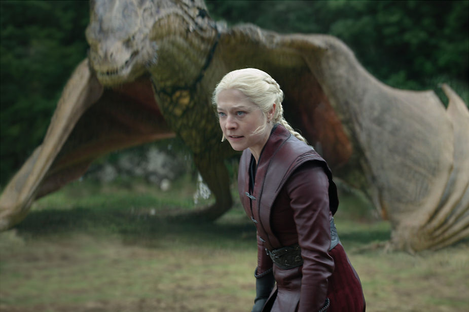 Blonde woman in maroon leather stands tense before a dragon in a wooded clearing, with a fierce, dangerous mood
