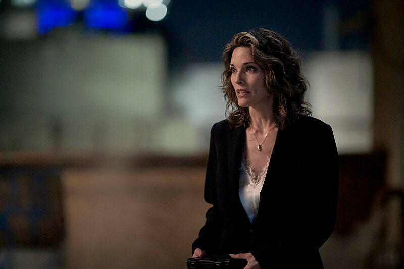 Woman in a black blazer holds a camera indoors, looking off to the side against a blurred, softly lit background.