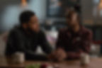 Two somber man and woman sit at a table holding hands, with white mugs, her maroon Nike jacket in a dim room.