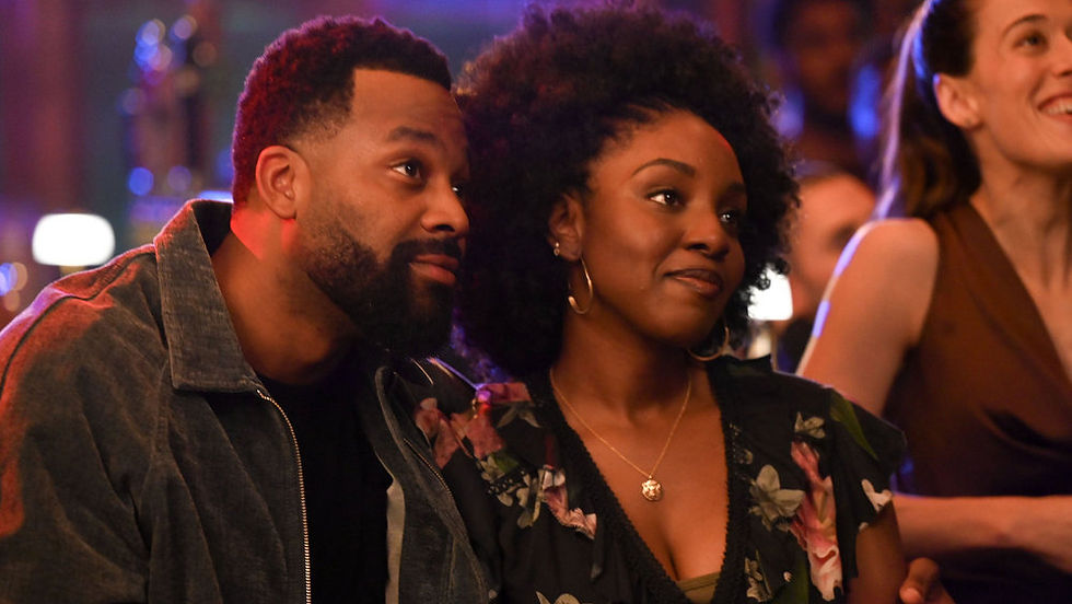 Smiling couple seated at a warmly lit event, watching something offscreen with a blurred crowd in the background.
