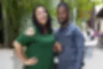 Smiling couple holding hands, standing outdoors with a leafy background. Woman in green, man in denim. Bright, cheerful atmosphere.