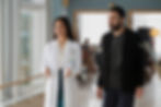 Female doctor in a white lab coat talks beside a bearded man in a hospital hallway, with bright windows and offices behind them