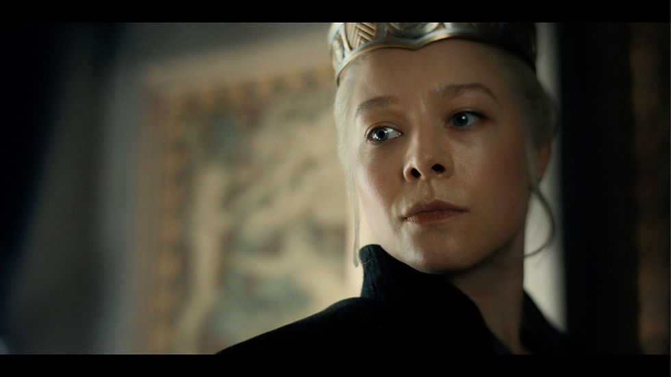 Person with a gold crown gazes to the side, wearing a dark outfit. The blurred background features a framed artwork, creating a regal mood.