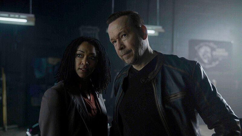 Two serious people stand in a dim industrial room with overhead lights, facing the camera in dark clothing.