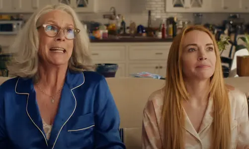 Two women in pajamas sit on a couch in a kitchen. One looks surprised in blue, the other calm in pink. White cabinets in the background.