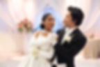 Bride and groom dance closely in a softly lit wedding hall with white drapes and pink flowers, sharing a romantic gaze.