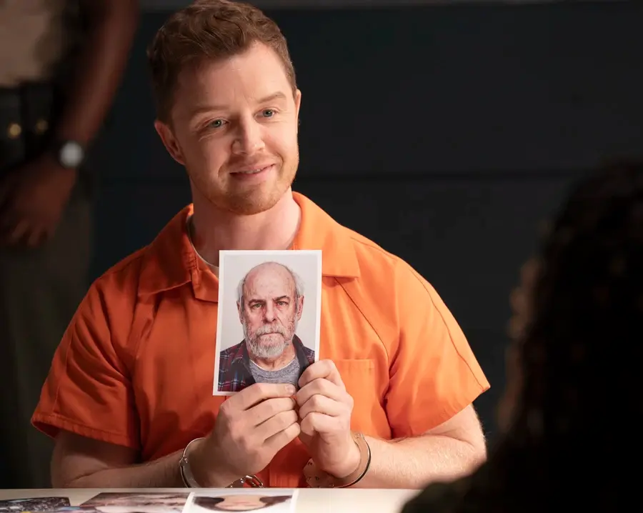 A man in an orange prison uniform, smiling and holding a mugshot photo of an older bearded man in a plaid shirt. Dark background.