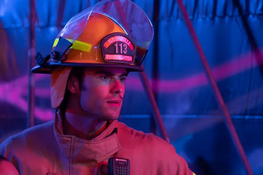 Firefighter in full gear looking focused under blue and pink lights. Helmet reads "Nashville Firefighter 113." Background has a tent-like structure.