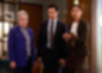 Three people in an office look concerned. An elderly woman in a blue blazer stands beside a man in a suit and a woman holding a notepad.