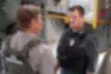 Two men in tactical gear stand in an industrial setting. One wears a "U.S. Marshal" vest. The mood appears tense. Equipment is visible.