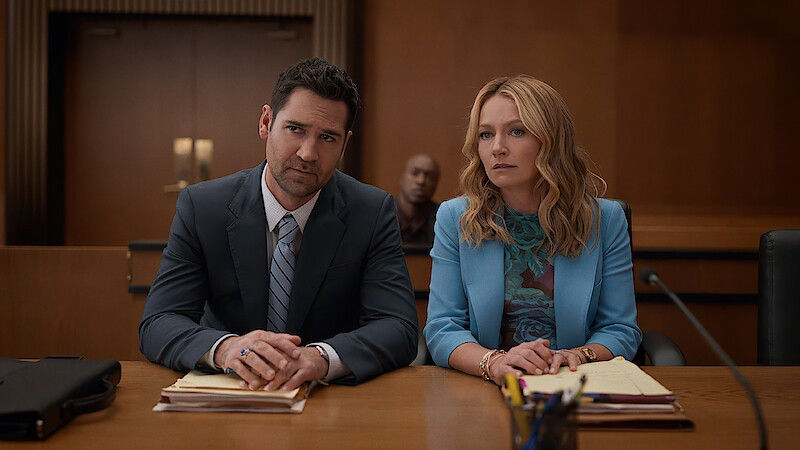 Two people sit at a courtroom table, appearing serious. Both are in suits, with papers and a briefcase in front. Wood-paneled background.