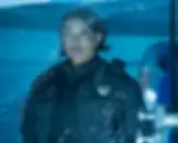 A police officer in uniform stands behind a sheet of plastic, looking serious. The background is blue, adding a cold, tense mood.