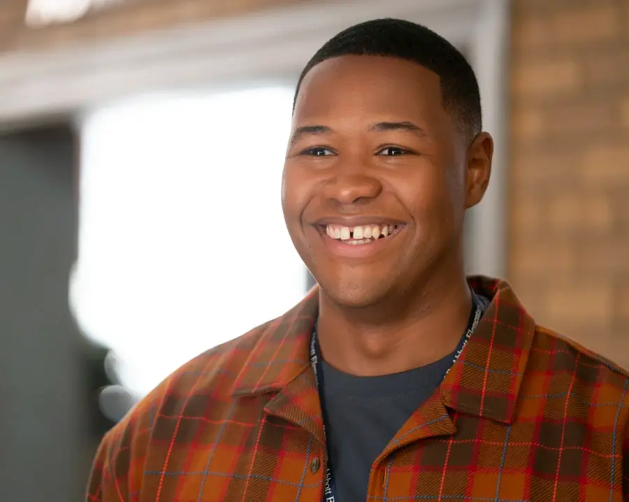 Smiling person in a plaid shirt stands indoors against a blurred brown brick wall, exuding a cheerful and friendly mood.