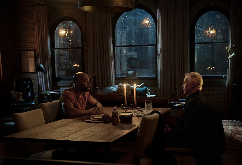 Two men sit at a candlelit table in a dimly lit room with arched windows showing a rainy cityscape outside, creating a tense atmosphere.