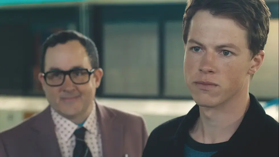 Serious young man in foreground with older man behind him in an indoor setting, both looking forward.