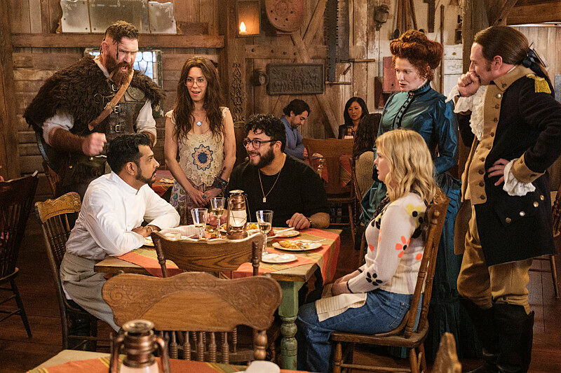 A group of people, some in period costumes, engaged in lively conversation at a rustic wooden table. Warm lighting creates a cozy atmosphere.