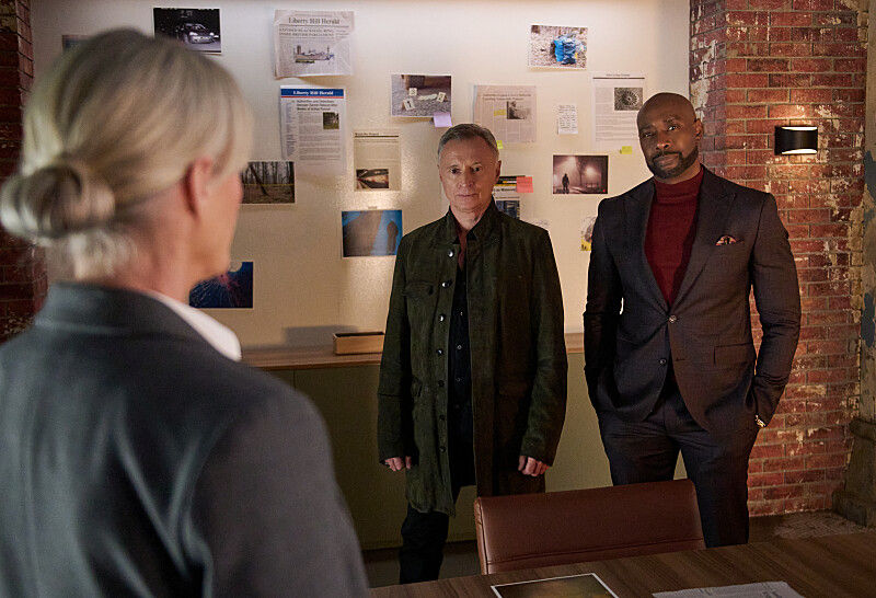 Two men stand facing a woman in an office with a board covered in papers and photos. Exposed brick walls add a vintage feel.