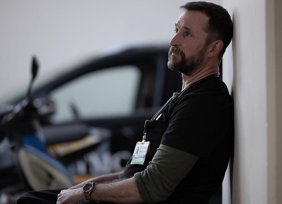 Bearded man in black shirt, wearing a "Doctor" badge, leans against a wall pensively. A car is blurred in the background.