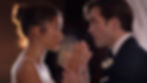 A man and woman in formal attire hold hands, gazing at each other intensely. Soft lighting highlights them against a blurred background.