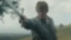 A determined person extends a sword forward in a grassy landscape, wearing a grey shirt. The cloudy sky adds a dramatic mood.