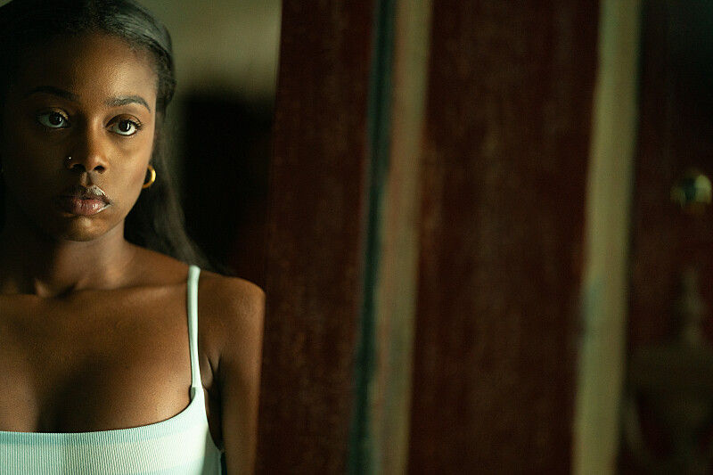 Close-up of a serious young Black woman in a white tank top, standing indoors beside a wooden mirror frame.