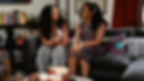 Two women sit on a black couch chatting and smiling in a cozy living room with bookshelves, patterned pillows, and a coffee table.