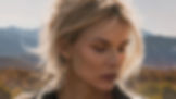 Woman with blonde hair looks down pensively against a mountainous backdrop. Sunlight highlights her face and leather jacket.