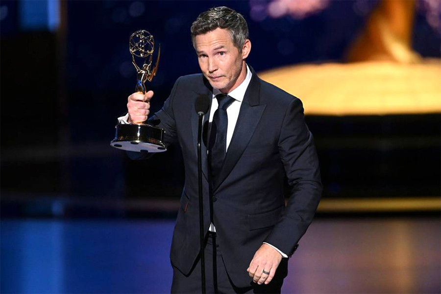 Man in a suit holds a trophy and speaks into a microphone on stage. The background is blue and gold, suggesting an awards ceremony.