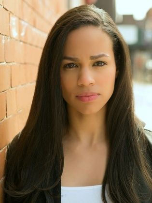 Woman with long hair stands against a brick wall, looking serious. Sunlight casts shadows, creating a warm and contemplative mood.