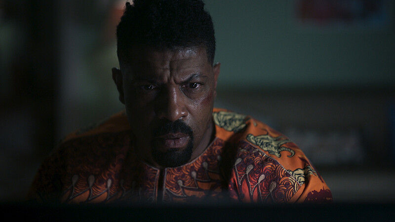 Serious man in orange patterned shirt stares forward in a dim, moody room.