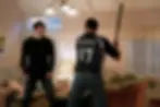 Two men in a living room, one holding a bat wearing a "RIKER 17" shirt. The other looks alarmed. Cozy decor, warm lighting. Tense mood.